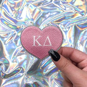 May include: Pink heart-shaped patch with silver glitter trim and white embroidered letters "KA" in the center.