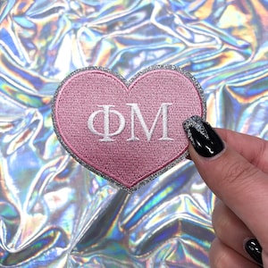 May include: Pink heart-shaped patch with a silver glitter border and white embroidered letters "ΦΜ".