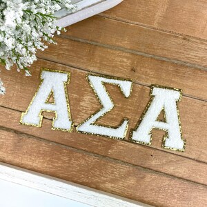 May include: White felt letters with gold glitter outlining spelling out "AZA" on a wood background.