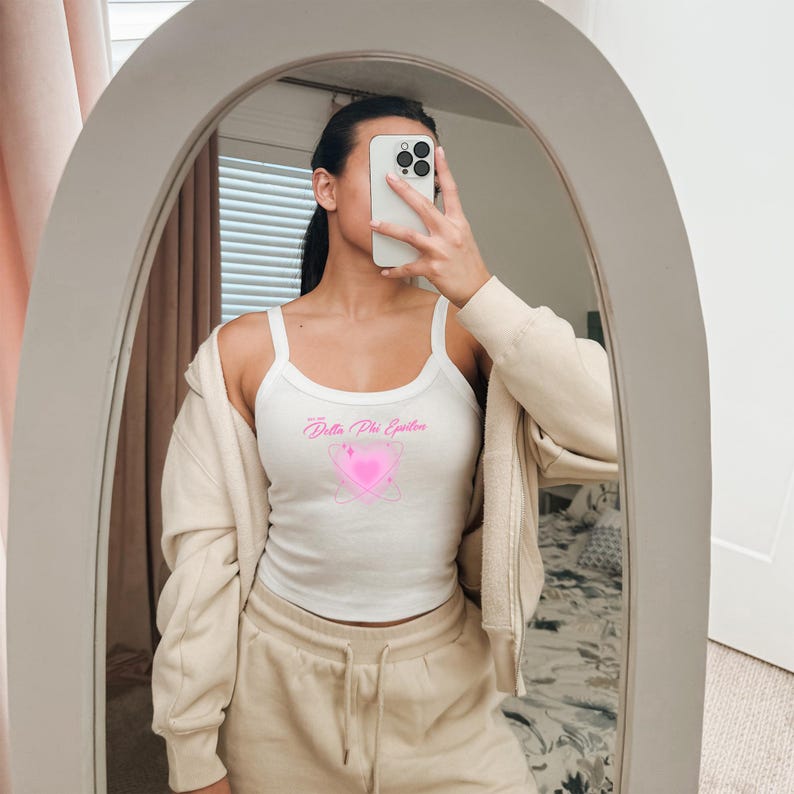 Puede incluir: Camiseta sin mangas blanca con el texto rosa "Delta Phi Epsilon" y un dise&ntilde;o de coraz&oacute;n rosa. La camiseta es de estilo acanalado.