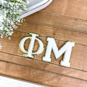 May include: White felt letters spelling "ΦΜ" with gold glitter outlining the letters. The letters are on a wood surface.