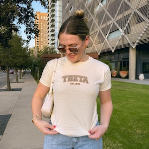 May include: A cream-colored t-shirt with the word "Theta" in a brown leopard print design, with "EST. 1870" printed below. The shirt is short-sleeved and has a crew neck.