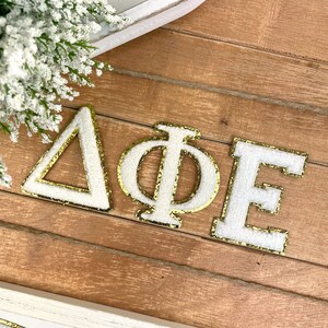 May include: White felt letters with gold glitter trim spelling out "Delta Phi Epsilon" on a wooden surface.