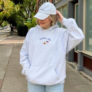 May include: A gray hooded sweatshirt with the text "ALPHA PHI" and a small embroidered bear and American flag. The model is wearing a white baseball cap with the text "undisclosed" on the front.