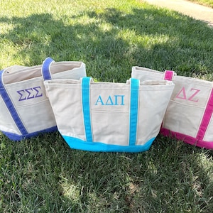 May include: Three canvas tote bags with Greek letters embroidered on the front. The bags have a natural beige color with accents in blue and pink. The bags are sitting on green grass.