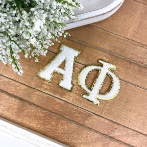 May include: White felt letters spelling "A".  A gold glitter felt letter "Phi" is next to the "A".  The letters are on a wood surface.