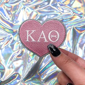 May include: Pink heart-shaped patch with silver glitter trim and the letters "KAO" in white embroidery.