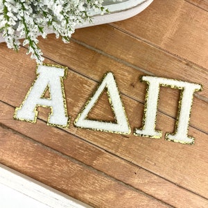 May include: White and gold Greek letter patches spelling out the letters A, Δ, and Π. The letters are white with a gold glitter border. The patches are on a wooden surface.