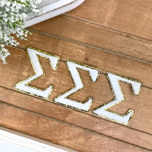 May include: Three white felt letters with gold glitter trim spelling out the Greek letters "ΣΣΣ".