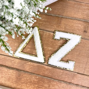 May include: White felt letters with gold glitter trim spelling out "ΔZ" on a wood background.