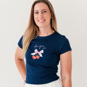 May include: A navy blue t-shirt with a pink bow and two red cherries. The shirt has the text "die que" and "xoxo" printed on it.