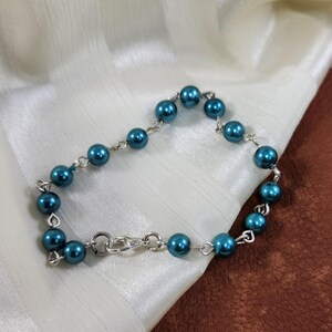 May include: A teal blue beaded bracelet with a silver clasp. The bracelet is made of small, round beads that are strung together on a silver chain.