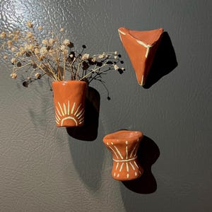 May include: Three terracotta-colored ceramic magnets with white painted designs. One is a small vase holding dried flowers, another is a triangular shape, and the third is a small, rounded shape. The magnets are attached to a gray surface.