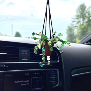 Peut inclure: Un charm de voiture suspendu avec un petit pot marron contenant une plante en perles. La plante est ornée de perles vertes, jaunes et roses, suspendue par une ficelle noire. Le charme est suspendu devant le tableau de bord d'une voiture.