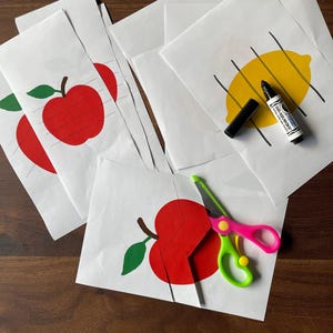 Op de afbeelding: Witte papieren vellen met rode appel- en gele citroenillustraties, een zwarte stift en roze en groene schaar. De appel- en citroenontwerpen hebben stippellijnen om te knippen.