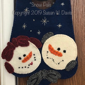 May include: A blue felt door hanger with two snowmen, one with a red bow. The snowmen are embroidered with black eyes, orange noses, and red mouths. The door hanger has white embroidered stars and the text "Snow Pals" and "Copyright 2019 Susan W. Davis".