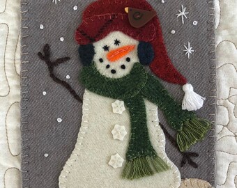 May include: A felt snowman with a red hat, green scarf, and black boots. The snowman is standing in a snowy scene with white snowflakes.