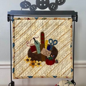 May include: A small quilted wall hanging with a brown basket filled with sewing supplies, including scissors, thread, and a tape measure. The basket is surrounded by sunflowers and red apples. The wall hanging is displayed on a black metal stand with a decorative top featuring a needle and buttons.