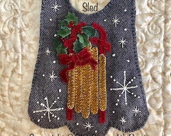 May include: A grey felt Christmas ornament featuring a red sled and holly design. The ornament has a hole at the top for hanging and the text "My Red Sled" is embroidered on the front. Copyright 2019 Susan W. Davis.