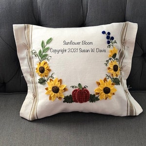 May include: A white pillow with a brown and white striped border. The pillow is decorated with embroidered sunflowers, leaves, and a pumpkin. The text "Sunflower Bloom Copyright 2021 Susan W. Davis" is embroidered on the pillow.