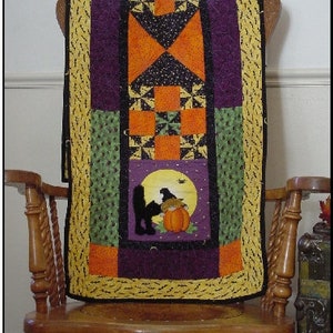 Op de afbeelding: Een Halloween-thema quilted tafelrunner met een ontwerp van een zwarte kat, pompoen en maan. De runner is gemaakt van verschillende stoffen in oranje, groen, paars en geel.