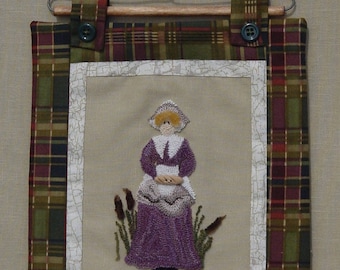 May include: A framed needlepoint wall hanging with a plaid border. The needlepoint depicts a woman in a purple dress standing in a field of tall grass.