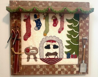 May include: A framed Christmas-themed wall hanging with a rustic aesthetic. It features a garland, stockings, a Christmas tree, and a fireplace scene. A pair of skis and a small shovel are attached to the frame. The color palette includes reds, greens, and browns.