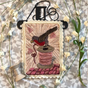 May include: A needlepoint embroidery of a robin perched on a spool of thread. The robin is red, gray, and brown, and the thread is pink. The embroidery is framed with a white scalloped border.