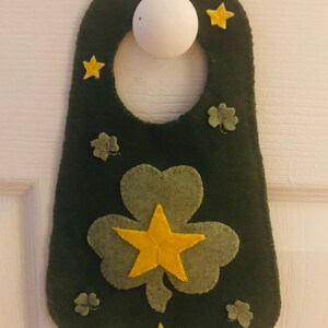 Puede incluir: Un colgador de puerta de fieltro verde con un diseño de trébol y estrellas doradas. El trébol está en el centro del colgador y tiene una estrella dorada en el medio. Hay tréboles más pequeños y estrellas doradas alrededor del trébol.