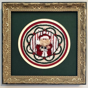 May include: A framed needlepoint artwork featuring a Santa Claus figure in a red and white striped suit holding a small gift. The artwork is surrounded by a green wreath with red berries and a white background.