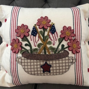 K&ouml;nnte beinhalten: Ein wei&szlig;es Kissen mit roten und blauen Streifen und einem roten Stern. Das Kissen hat einen braunen und wei&szlig;en karierten Topf mit roten Blumen und amerikanischen Flaggen.