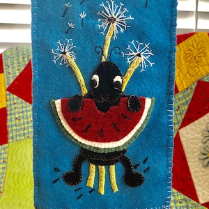 May include: A felt appliqué of a black ant holding a slice of watermelon with yellow dandelion stems and white seeds. The ant has large black eyes and is standing on a blue background.