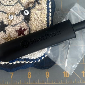 May include: A black "Smart Punch" tool with a long, cylindrical handle and a needle tip, alongside a clear plastic bag containing two needles. The tool rests on a patterned rug with star and letter details.