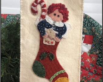 May include: A cream felt Christmas stocking with a red stocking applique. The stocking features an angel holding a candy cane and a tag that says "From Me". The angel has red hair and is wearing a blue dress.