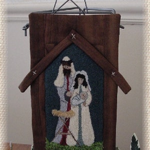 Op de afbeelding: Een rustieke kerstversiering met een houten structuur en een ster. Het midden toont een kerststal met Maria, Jozef en het kindje Jezus. Het kunstwerk is omlijst door een bruine houten huisvorm met een ster.