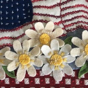 May include: A close-up of a handmade felt American flag quilt block with a red, white, and blue color scheme. The flag is decorated with white daisies with yellow centers. The daisies are arranged in a row at the bottom of the block.