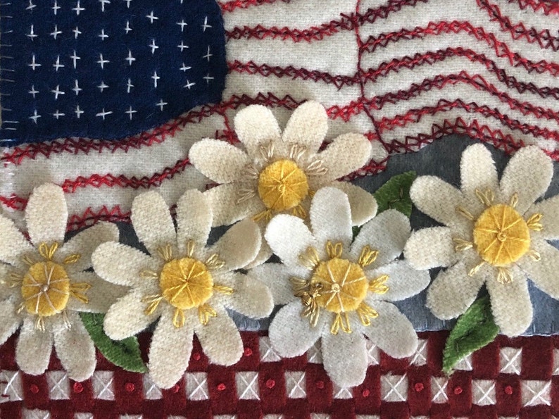 May include: A close-up of a handmade felt American flag quilt block with a red, white, and blue color scheme. The flag is decorated with white daisies with yellow centers. The daisies are arranged in a row at the bottom of the block.