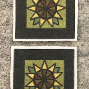 May include: Two square fabric coasters with a black border and a green background. Each coaster features a stylized starburst design with yellow, brown, and green accents.