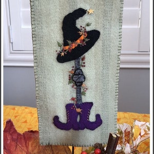 May include: A decorative, rectangular wall hanging featuring a black witch's hat with a gold star and autumn foliage. Below the hat is a stem with a decorative element and purple witch boots. The background is a light green fabric with a stitched border.