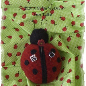 May include: A red and black ladybug pincushion with black dots and a black stripe. The pincushion is sitting on a green fabric with a pattern of red ladybugs.