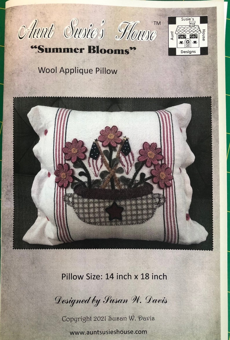 K&ouml;nnte beinhalten: Ein wei&szlig;es Woll-Applikationskissen mit einem Design aus einem Blumenkorb und Flaggen. Das Kissen ist 14 Zoll mal 18 Zoll gro&szlig;. Der Text "Summer Blooms" ist auf der Vorderseite des Kissens aufgedruckt.