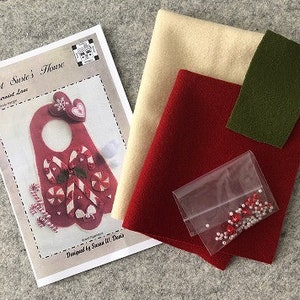 May include: A felt craft kit for making a peppermint door hanger. The kit includes red, white, and green felt, a bag of red and white beads, and instructions. The instructions include the title "Aunt Susie's House" and the words "Peppermint Love".