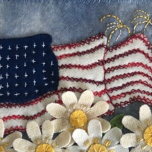 May include: A patriotic quilt block featuring a red, white, and blue American flag with white stars, red and white stripes, and yellow embroidered fireworks. The flag is surrounded by white daisies with yellow centers.