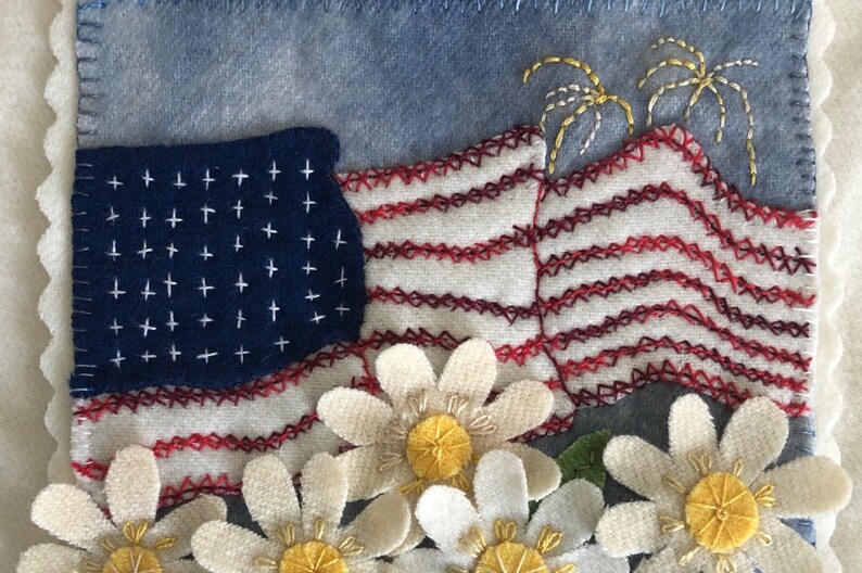 May include: A patriotic quilt block featuring a red, white, and blue American flag with white stars, red and white stripes, and yellow embroidered fireworks. The flag is surrounded by white daisies with yellow centers.