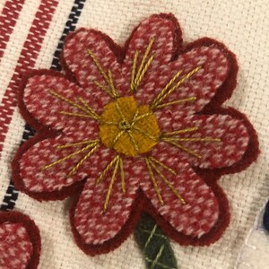 K&ouml;nnte beinhalten: Eine rote Filzblume mit gelber Mitte und goldener Stickerei. Die Blume ist auf einen wei&szlig;en Stoffhintergrund gestickt.
