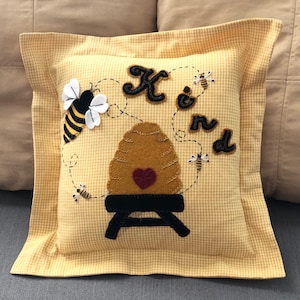 May include: Yellow and white gingham pillow with a beehive, bees, and the word "Kind" embroidered on it.