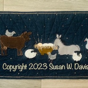 May include: A blue quilted wall hanging with a nativity scene. The scene includes a cow, a donkey, a manger with a baby, and a flock of white sheep. The text "Copyright 2023 Susan W. Davis" is at the bottom of the hanging.