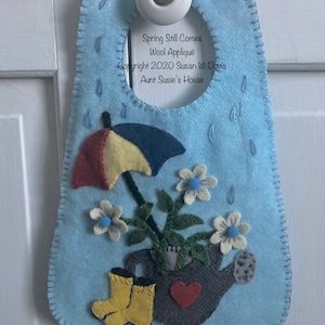 May include: A blue felt door hanger with a watering can, flowers, and a yellow umbrella. The text "Spring Still Comes Wool Applique Copyright 2020 Susan W. Davis Aunt Susie's House" is on the hanger.