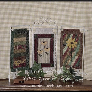 May include: Three small quilted wall hangings with a sunflower theme displayed on a white metal rack. The hangings are made with a variety of fabrics in shades of green, brown, red, and yellow. The text "Sunflower Quilts" and "©2008 Susan W. Davis" is visible below the hangings. The website address "www.auntsusieshouse.com" is visible below the text.