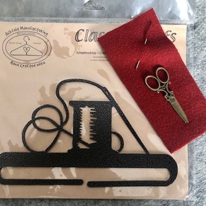 May include: A black metal wire hanger shaped like a sewing machine with a spool of thread and a needle. The hanger is packaged with a red felt square, a pair of small scissors, and two needles. The package is labeled "Classic Crafts" and "Ack-Feld Manufacturing Established 1990 Hand Crafted Wire".